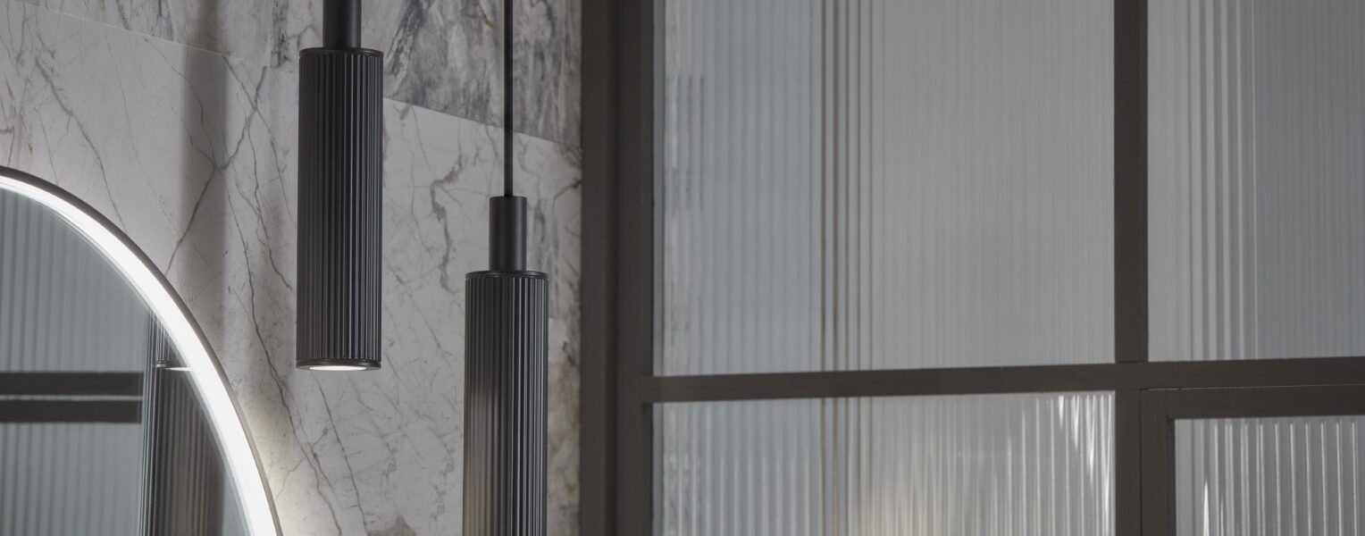 The primary focus is two pendant lights, which are identical in design. They are long, cylindrical, and appear to be made of a dark-coloured metal, possibly with a textured or ribbed surface. They hang from thin cables, suggesting a minimalist and contemporary style. The lights are positioned at slightly different heights, adding visual interest.