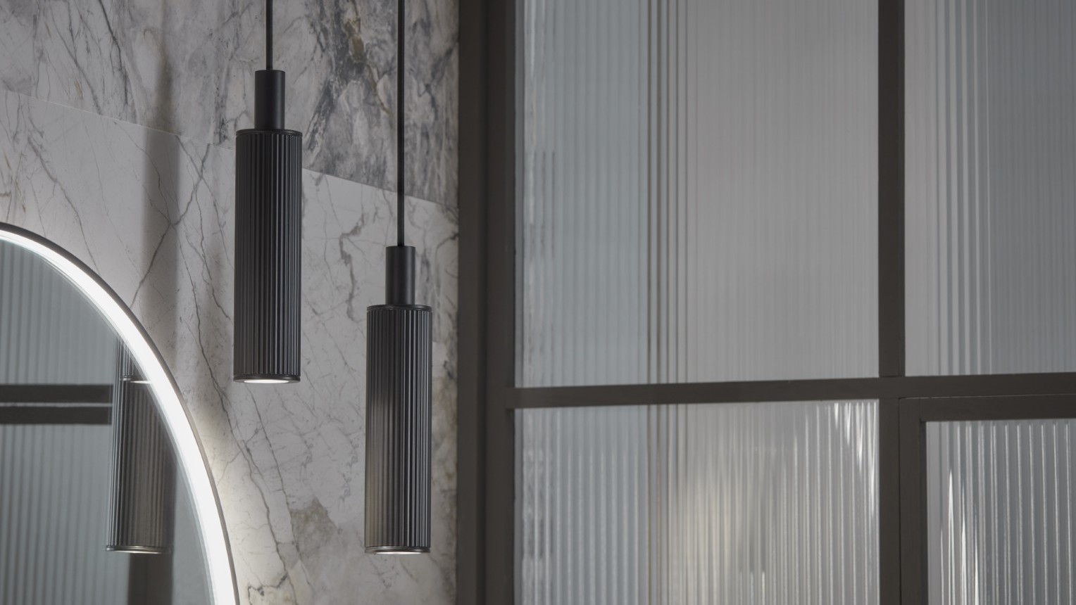 The primary focus is two pendant lights, which are identical in design. They are long, cylindrical, and appear to be made of a dark-coloured metal, possibly with a textured or ribbed surface. They hang from thin cables, suggesting a minimalist and contemporary style. The lights are positioned at slightly different heights, adding visual interest.