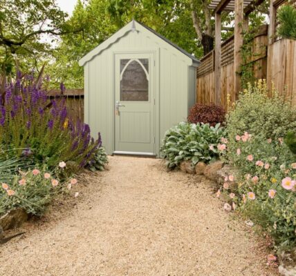 A light green wooden shed with a peaked roof and a white, arched windowed door at the far end.