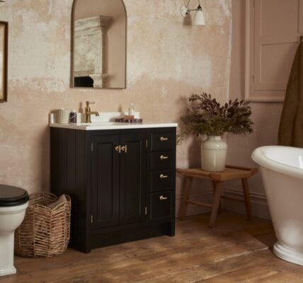 A bathroom with a rustic, vintage aesthetic. The walls have a textured, plaster-like finish in a light pinkish-beige hue. A dark wood vanity with a white countertop and gold hardware is centred in the frame