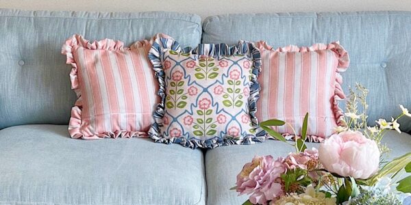 A close-up shot of a light blue tufted couch with three decorative pillows. The two outer pillows feature pink and white vertical stripes with ruffled edges. The centre pillow has a floral pattern with pink flowers and blue accents on a white background, also with ruffled edges.