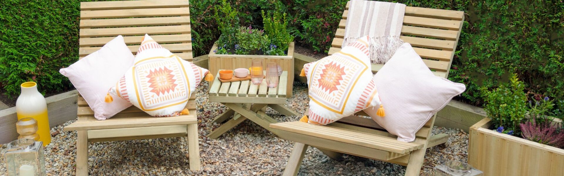 Two slatted wooden chairs and a small slatted wooden table arranged on a gravel surface. The chairs have cushions on them, and there are a few decorative items on the table, along with lanterns on the ground nearby.