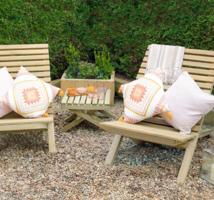 Two slatted wooden chairs and a small slatted wooden table arranged on a gravel surface. The chairs have cushions on them, and there are a few decorative items on the table, along with lanterns on the ground nearby.