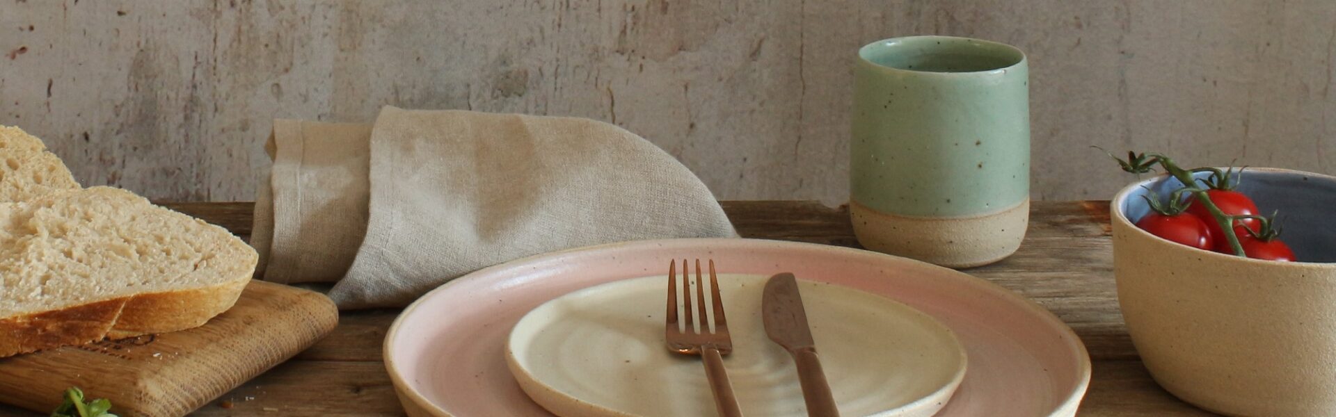 A stack of two ceramic plates. The larger plate has a light beige exterior and a soft pink interior. The smaller plate is a creamy white. A fork and knife rest on the smaller plate.