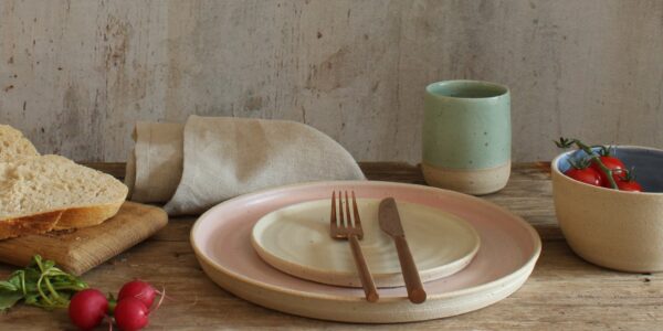 A stack of two ceramic plates. The larger plate has a light beige exterior and a soft pink interior. The smaller plate is a creamy white. A fork and knife rest on the smaller plate.