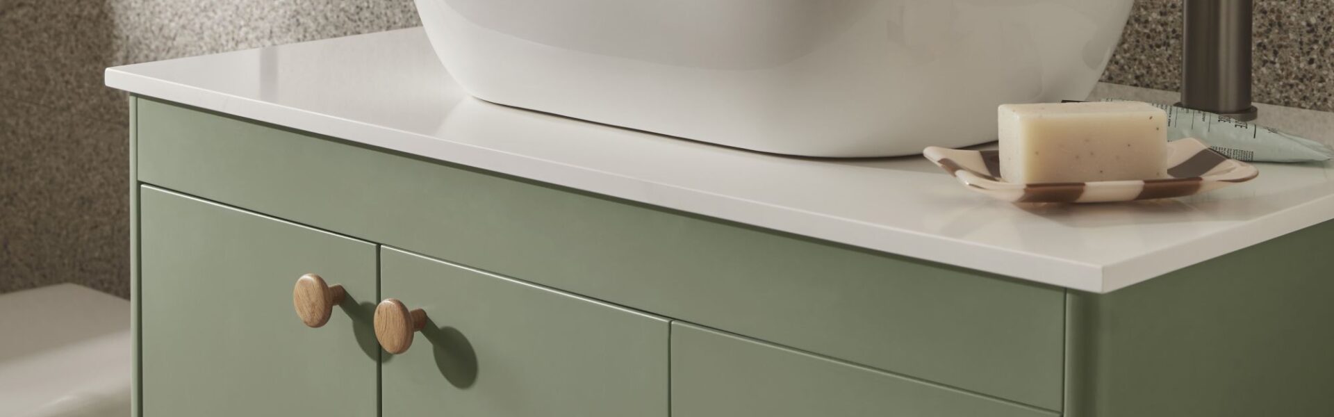 The image shows a close-up of a bathroom vanity unit. The vanity has a modern, minimalist design with a light sage green cabinet and a white countertop. A sleek, white, oval-shaped countertop basin sits on top of the counter.