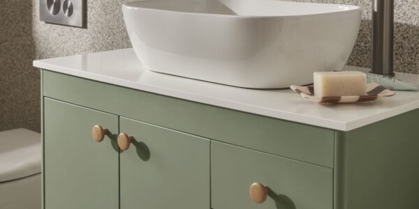 The image shows a close-up of a bathroom vanity unit. The vanity has a modern, minimalist design with a light sage green cabinet and a white countertop. A sleek, white, oval-shaped countertop basin sits on top of the counter.