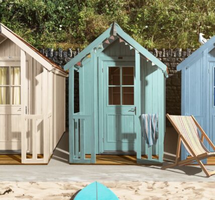 Three colourful beach hut-style sheds on a sandy surface, with surfboards and a beach chair nearby