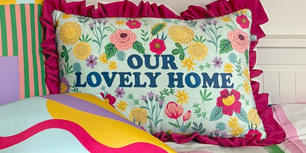 Close-up of a decorative pillow on a bed featuring a floral design in pink, yellow, and green on a light blue background, with the phrase ‘Our Lovely Home’ prominently displayed