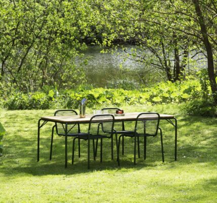 A vibrant outdoor scene features a wooden rectangular dining table with four black metal chairs, set on a lush green lawn.