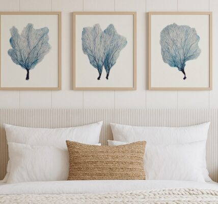 A bedroom scene features a large bed with a white pleated headboard and white bedding. A textured brown throw pillow rests on the bed. Above the bed, a set of three framed art prints, each depicting blue coral, are hung on a white panelled wall.