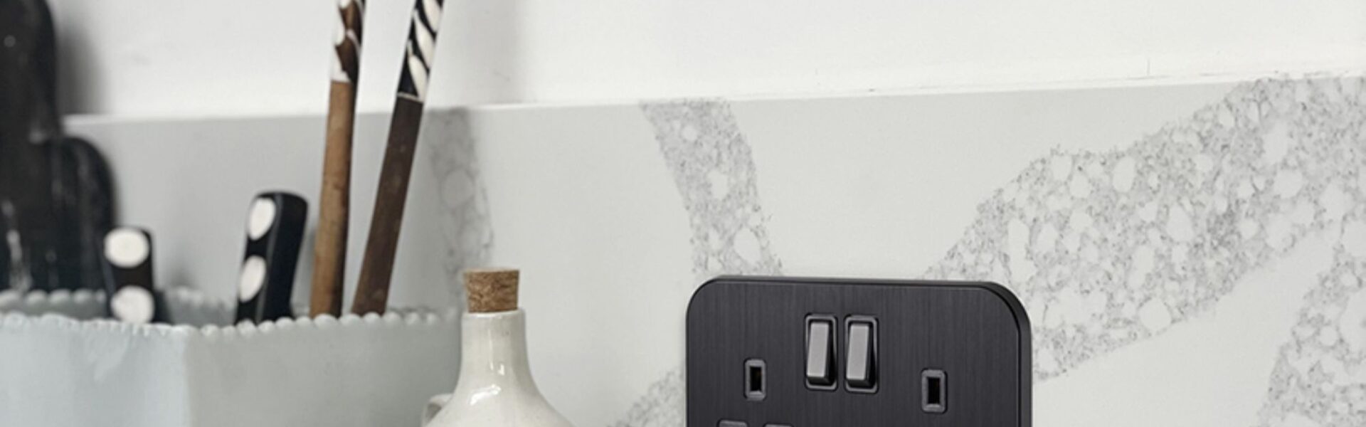 A sleek, black double UK plug socket (13A) is mounted on a grey and white marble-effect splashback in a modern kitchen.