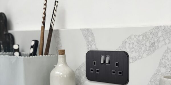 A sleek, black double UK plug socket (13A) is mounted on a grey and white marble-effect splashback in a modern kitchen.