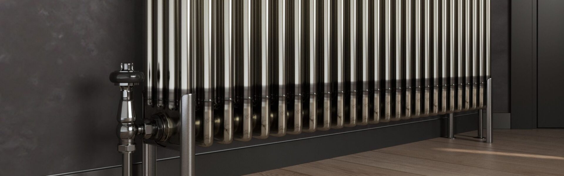 A bare metal, multi-column radiator stands on a light wooden floor with dark grey skirting boards and a dark grey wall behind it. To the left, a silver valve and pipe connect to the radiator, and on the right, two matching silver feet support it.