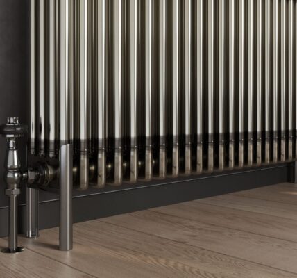 A bare metal, multi-column radiator stands on a light wooden floor with dark grey skirting boards and a dark grey wall behind it. To the left, a silver valve and pipe connect to the radiator, and on the right, two matching silver feet support it.