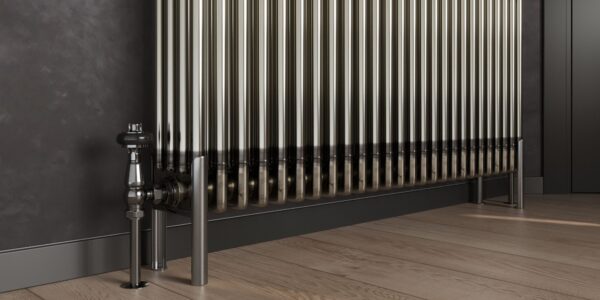 A bare metal, multi-column radiator stands on a light wooden floor with dark grey skirting boards and a dark grey wall behind it. To the left, a silver valve and pipe connect to the radiator, and on the right, two matching silver feet support it.