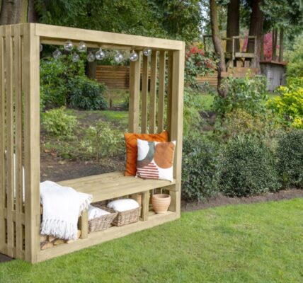 A natural wood-stained pergola with an integrated bench seat is positioned on a vibrant green lawn. The pergola has a rectangular shape with slatted sides and an open roof, from which hangs a string of decorative globe lights. On the bench, there are several cushions in warm tones of orange, brown, and cream.