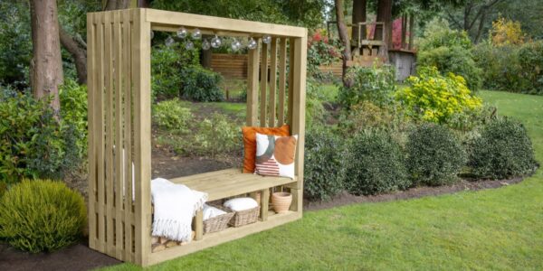 A natural wood-stained pergola with an integrated bench seat is positioned on a vibrant green lawn. The pergola has a rectangular shape with slatted sides and an open roof, from which hangs a string of decorative globe lights. On the bench, there are several cushions in warm tones of orange, brown, and cream.