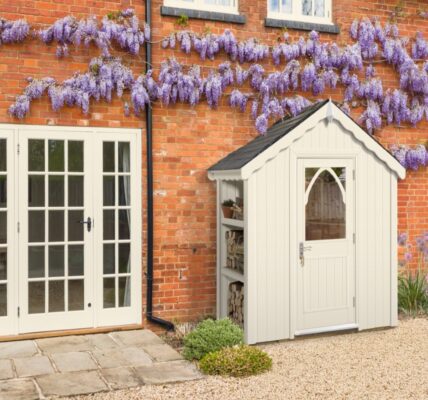 A quaint cream-coloured shed stands nestled beside a rustic brick house. The shed features a dark roof, a charming arched window in its door, and an open shelving unit on its left side, holding what appears to be firewood and a potted plant.