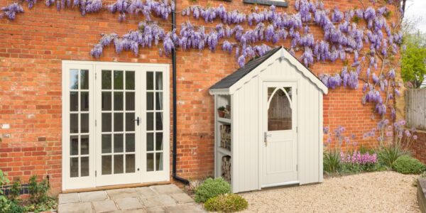 A quaint cream-coloured shed stands nestled beside a rustic brick house. The shed features a dark roof, a charming arched window in its door, and an open shelving unit on its left side, holding what appears to be firewood and a potted plant.