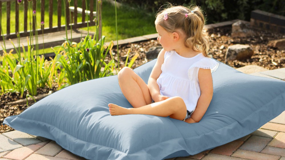 A young child sits comfortably on a large, seafoam blue bean bag cushion outdoors