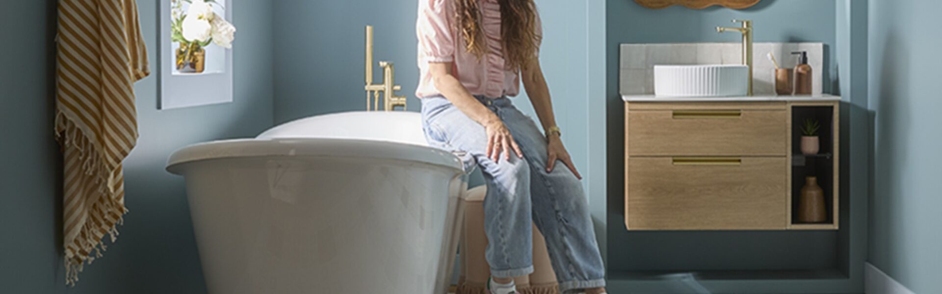 A woman with long, dark hair is seated on the edge of a white, freestanding bathtub in a bathroom. She is wearing a light-coloured, long-sleeved top and blue jeans. To her right, there is a wooden wall-mounted vanity unit with a white circular sink and a gold-coloured faucet.