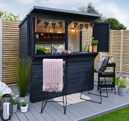 A garden bar made from a dark-painted wooden shed with a pent roof. The front opens up to reveal a bar area with shelves stocked with bottles, glasses, and bar tools. String lights are hung inside.