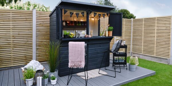 A garden bar made from a dark-painted wooden shed with a pent roof. The front opens up to reveal a bar area with shelves stocked with bottles, glasses, and bar tools. String lights are hung inside.