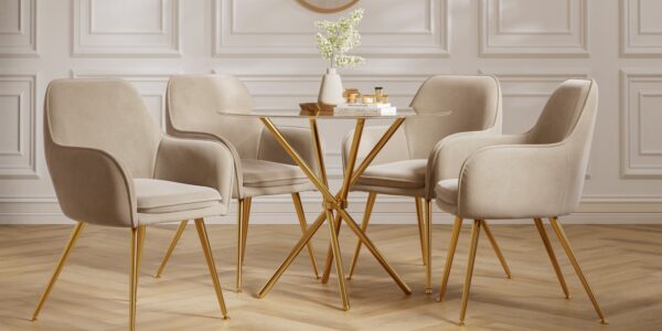 A round glass dining table with a gold base is surrounded by four beige velvet dining chairs with gold legs. The table is adorned with a white vase holding white flowers and a stack of books with some decorative items on top. The room features white panelled walls and a light-coloured wooden floor.