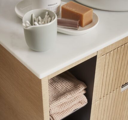 A close-up shot of a bathroom vanity unit. The unit has a light wood finish with a subtle fluted or ridged texture on the drawer fronts. It features a clean white countertop.