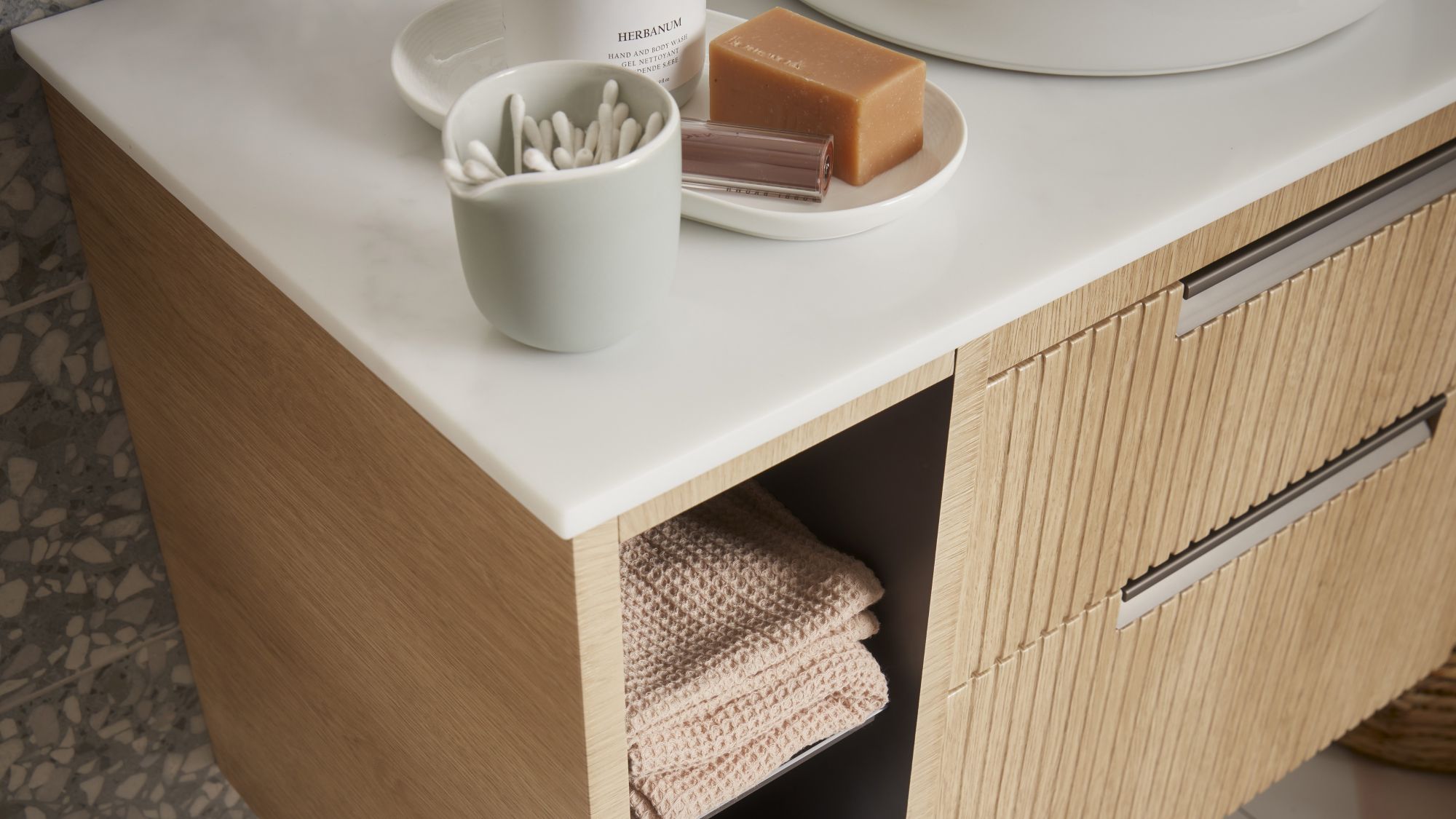 A close-up shot of a bathroom vanity unit. The unit has a light wood finish with a subtle fluted or ridged texture on the drawer fronts. It features a clean white countertop.