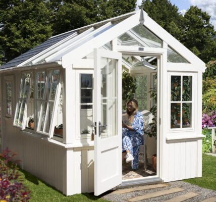 A charming, bright white greenhouse stands amidst a vibrant, sunlit garden. The greenhouse features a classic design with a gabled roof, a significant portion of which, along with the upper sections of its walls, is made of glass, allowing ample light to penetrate. The lower part of the greenhouse has solid, painted walls.