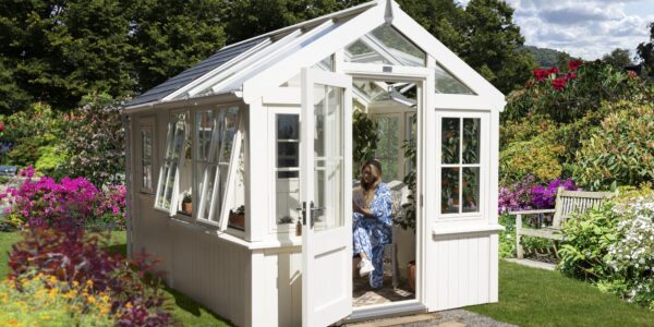 A charming, bright white greenhouse stands amidst a vibrant, sunlit garden. The greenhouse features a classic design with a gabled roof, a significant portion of which, along with the upper sections of its walls, is made of glass, allowing ample light to penetrate. The lower part of the greenhouse has solid, painted walls.