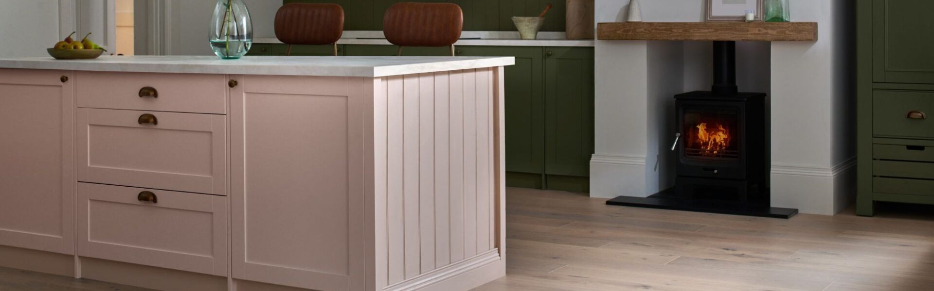 A beautifully styled kitchen features a large pink kitchen island with a white countertop and gold hardware, taking centre stage in the foreground. The floor throughout the kitchen is made of light-coloured engineered wood