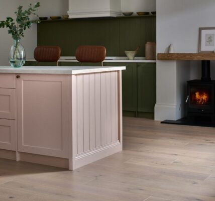 A beautifully styled kitchen features a large pink kitchen island with a white countertop and gold hardware, taking centre stage in the foreground. The floor throughout the kitchen is made of light-coloured engineered wood