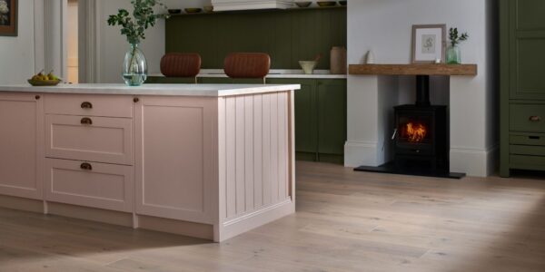 A beautifully styled kitchen features a large pink kitchen island with a white countertop and gold hardware, taking centre stage in the foreground. The floor throughout the kitchen is made of light-coloured engineered wood