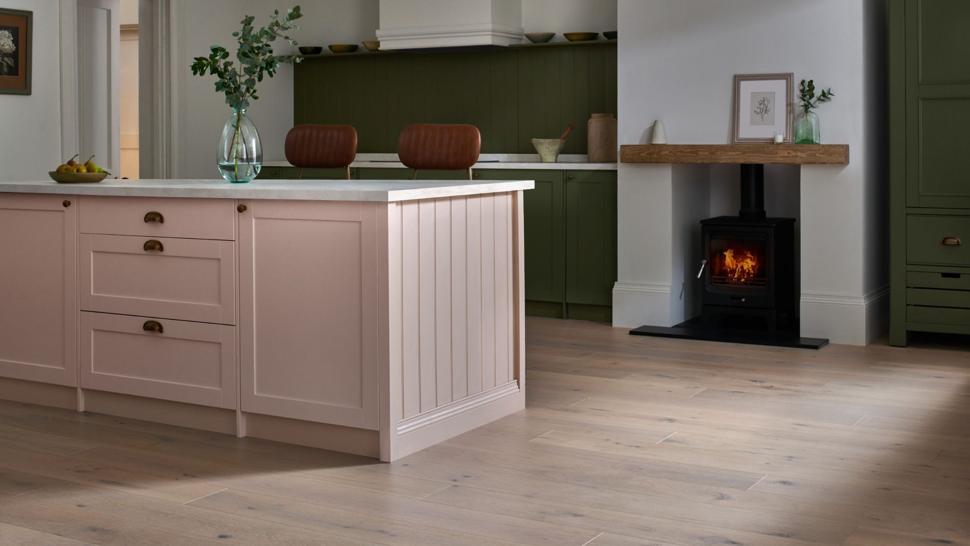 A beautifully styled kitchen features a large pink kitchen island with a white countertop and gold hardware, taking centre stage in the foreground. The floor throughout the kitchen is made of light-coloured engineered wood
