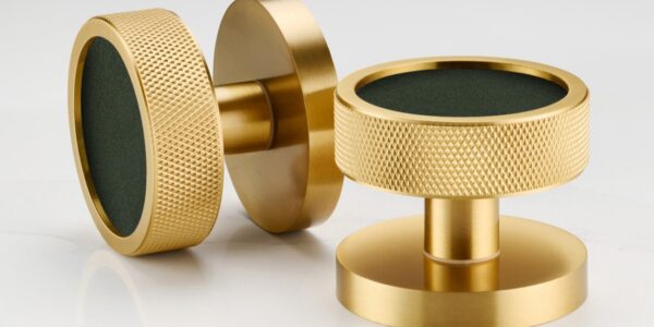 A close-up shot features a modern, round door knob in a striking design. The knob has a brass or gold-toned metallic outer ring, giving it a sleek and sophisticated look. The centre of the door knob is inlaid with what appears to be white terrazzo, speckled with various shapes and sizes of flecks in shades of black, grey, terracotta, and brown.