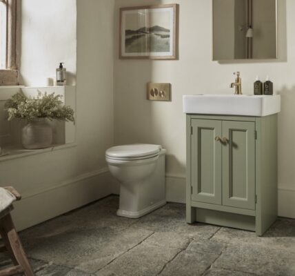 A warm and inviting bathroom features a Roper Rhodes Hampton 575 Freestanding Vanity Unit in Matt Sage Green with a white basin and gold-toned tap, positioned next to a white toilet. The vanity has two doors with round, light-coloured handles.