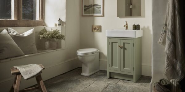 A warm and inviting bathroom features a Roper Rhodes Hampton 575 Freestanding Vanity Unit in Matt Sage Green with a white basin and gold-toned tap, positioned next to a white toilet. The vanity has two doors with round, light-coloured handles.