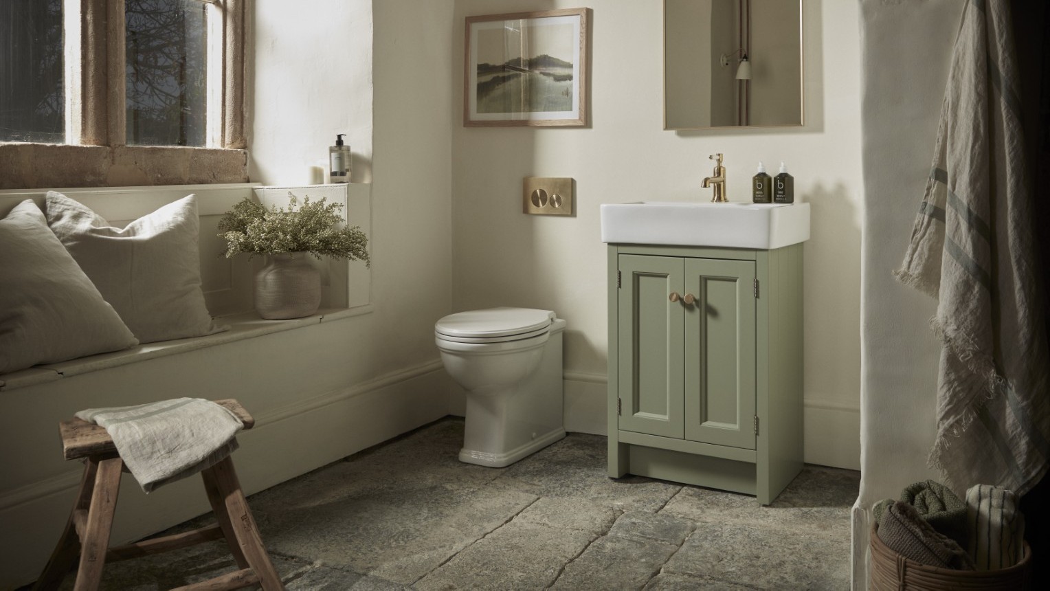 A warm and inviting bathroom features a Roper Rhodes Hampton 575 Freestanding Vanity Unit in Matt Sage Green with a white basin and gold-toned tap, positioned next to a white toilet. The vanity has two doors with round, light-coloured handles.