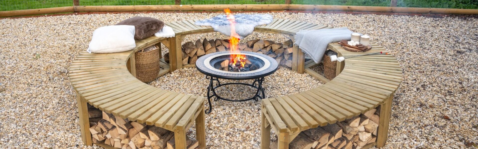 A low-angle shot shows a curved, wooden firepit bench, split into two semicircles, arranged around a central firepit. The benches are made of slatted wood and feature an under-bench shelf, which is filled with neatly stacked firewood. The benches are topped with soft cushions and blankets, suggesting a cosy and comfortable atmosphere.