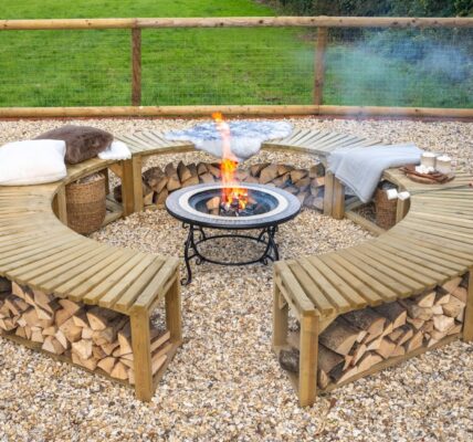 A low-angle shot shows a curved, wooden firepit bench, split into two semicircles, arranged around a central firepit. The benches are made of slatted wood and feature an under-bench shelf, which is filled with neatly stacked firewood. The benches are topped with soft cushions and blankets, suggesting a cosy and comfortable atmosphere.