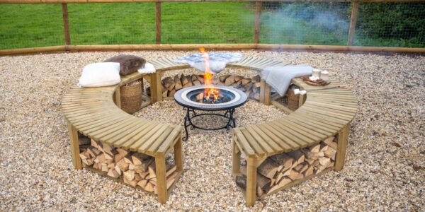 A low-angle shot shows a curved, wooden firepit bench, split into two semicircles, arranged around a central firepit. The benches are made of slatted wood and feature an under-bench shelf, which is filled with neatly stacked firewood. The benches are topped with soft cushions and blankets, suggesting a cosy and comfortable atmosphere.