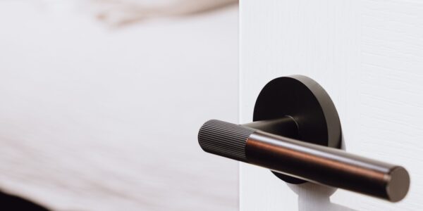A close-up shot of a modern brass linear lever handle in a bronze finish, installed on a white door. The handle has a sleek, cylindrical shape with fine ridging near the base, and it's mounted on a circular backplate.