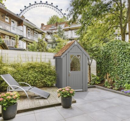 A stylish garden shed, named The Ludlow Shed by The Posh Shed Company, priced at £3999. The shed is grey with a red roof and features a window and a door, situated in a contemporary patio garden.