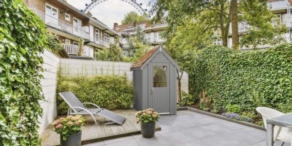 A stylish garden shed, named The Ludlow Shed by The Posh Shed Company, priced at £3999. The shed is grey with a red roof and features a window and a door, situated in a contemporary patio garden.