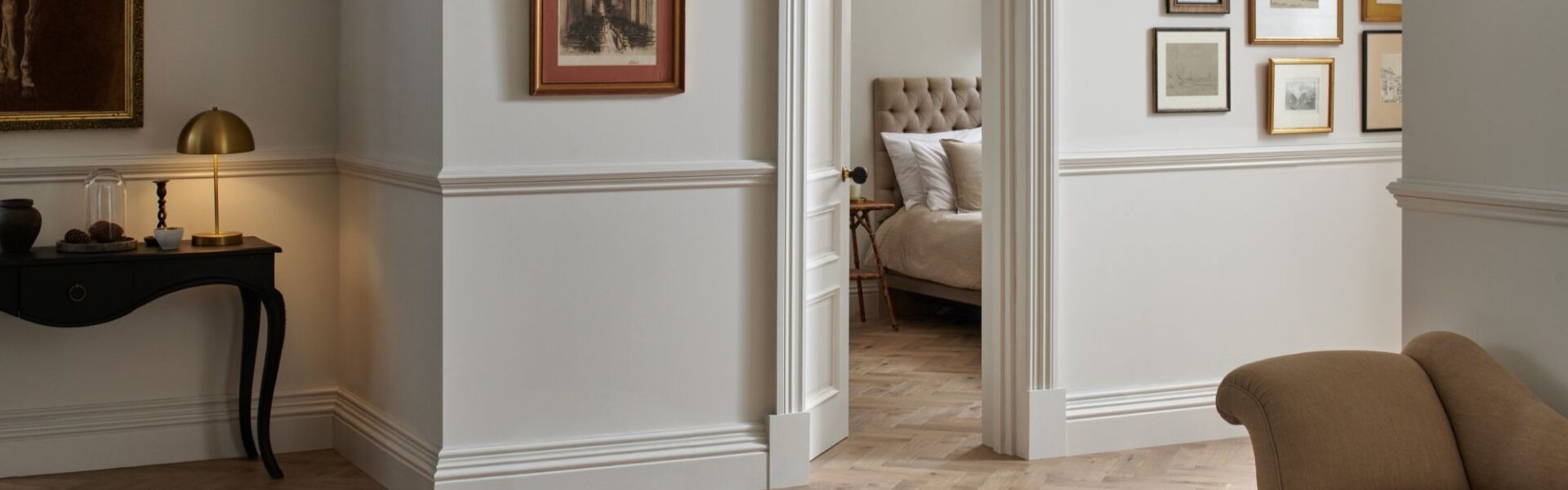 A stylish room featuring Woodpecker Flooring's Orkney Pewter Oak Herringbone Engineered Wood Flooring. The flooring is laid in a classic herringbone pattern, with varying shades of light brown and grey tones. The walls are painted in a light, neutral colour with white wainscoting and moulding.