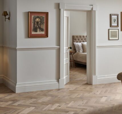 A stylish room featuring Woodpecker Flooring's Orkney Pewter Oak Herringbone Engineered Wood Flooring. The flooring is laid in a classic herringbone pattern, with varying shades of light brown and grey tones. The walls are painted in a light, neutral colour with white wainscoting and moulding.