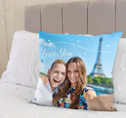 A custom photo cushion on a bed. The cushion features a picture of a smiling couple, with a man on the left embracing a woman on the right. Both are looking directly at the camera. Behind them, the Eiffel Tower is visible against a blue sky with some white decorative swirls and small hearts, and the words "Love You" written in a stylised font.